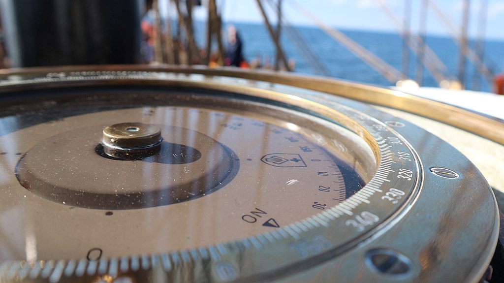 Close-up of a ship's compass, with a brass edge and clear navigational markings, set against a blurred view of the ocean and blue sky.