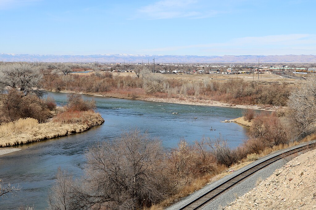 Wide river bends through barren landscape with dry shrubs and leafless trees. Train tracks run along the right. Distant town under clear sky. Calm, serene.