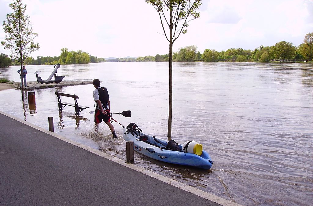 A person pulls a kayak onto a flooded riverside path. Water covers the walkway. Another person stands nearby, observing. Trees line the opposite riverbank.