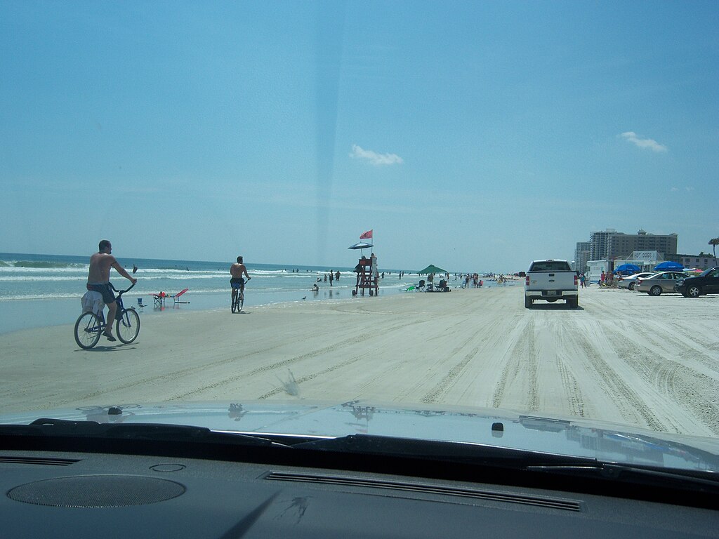 A sunny beach scene with people biking and walking along the sand. Cars drive nearby, and a lifeguard tower stands with a red flag. Blue sky overhead.