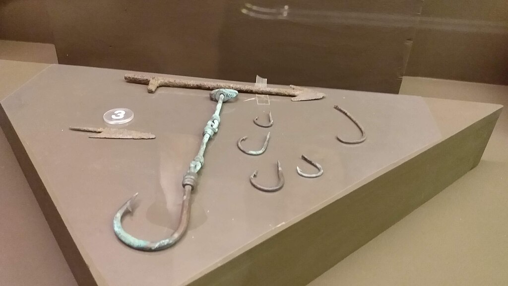 A display of various ancient fishing hooks of different sizes and shapes, made of metal, laid out on a triangular brown exhibit platform.