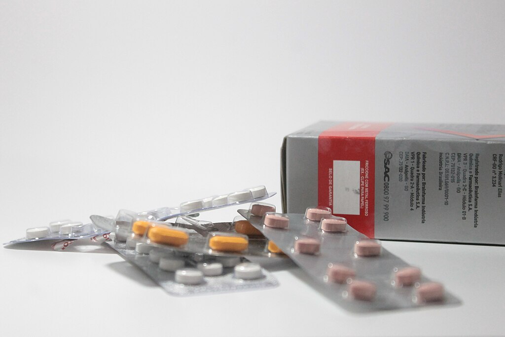 Blister packs with white, orange, and pink pills spill out of an opened grey box with red and white labeling, set against a plain white background.