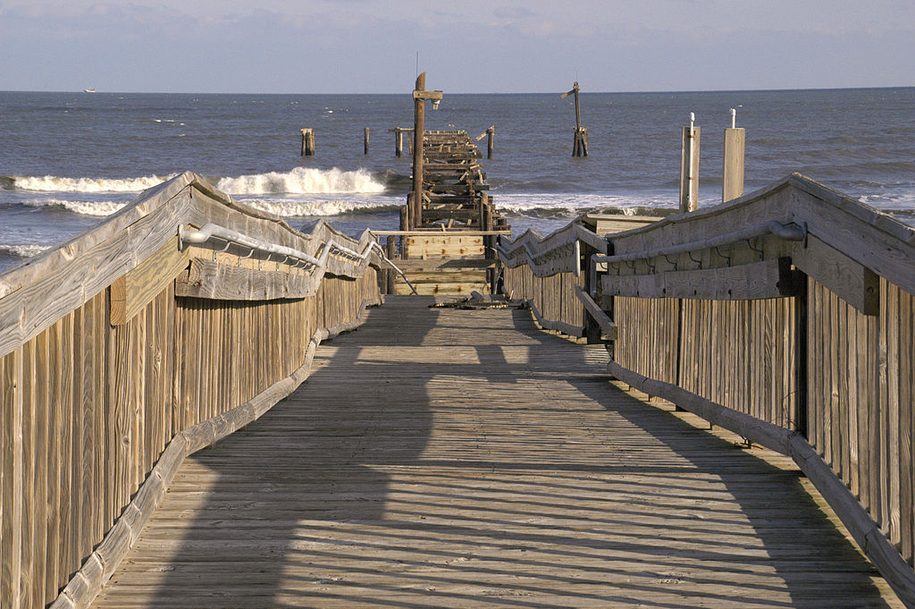 Wooden pier over the ocean, leading to a broken end, with gentle waves and a clear blue sky. A tranquil yet nostalgic atmosphere.