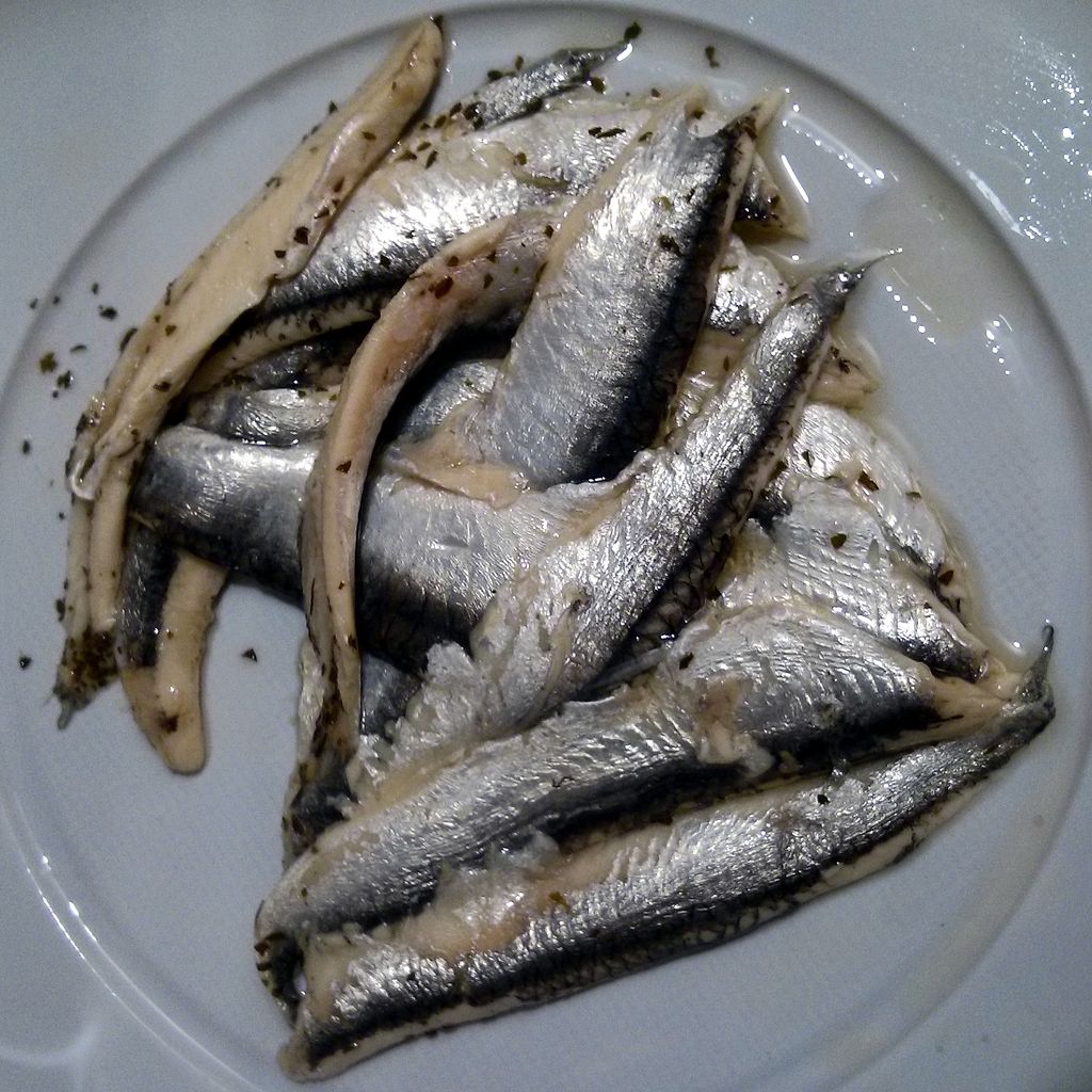 A white plate with shiny, marinated anchovy fillets arranged in a circular pattern, sprinkled with herbs, creating a fresh and appetizing look.