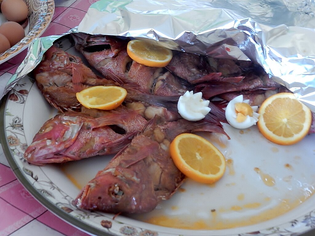 Baked fish on a floral-patterned platter, garnished with orange slices and halved boiled eggs, partially covered with foil, creating a rustic look.