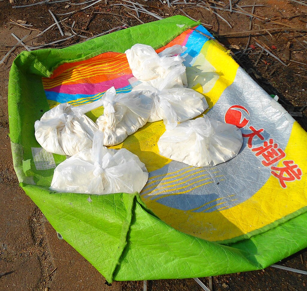 Plastic bags filled with white powder sit on a colorful, textured sack on sandy ground. The image suggests outdoor or market setting.