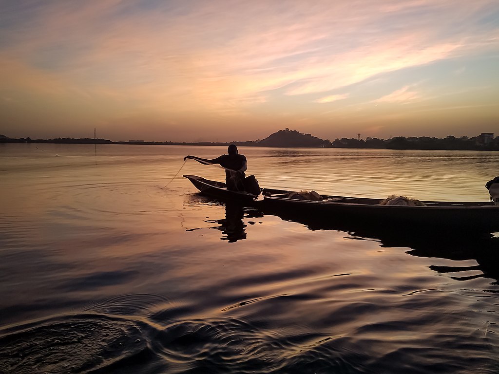 A silhouetted person fishes in a canoe on a calm lake during sunset. The sky is painted with warm orange and pink hues, creating a serene atmosphere.