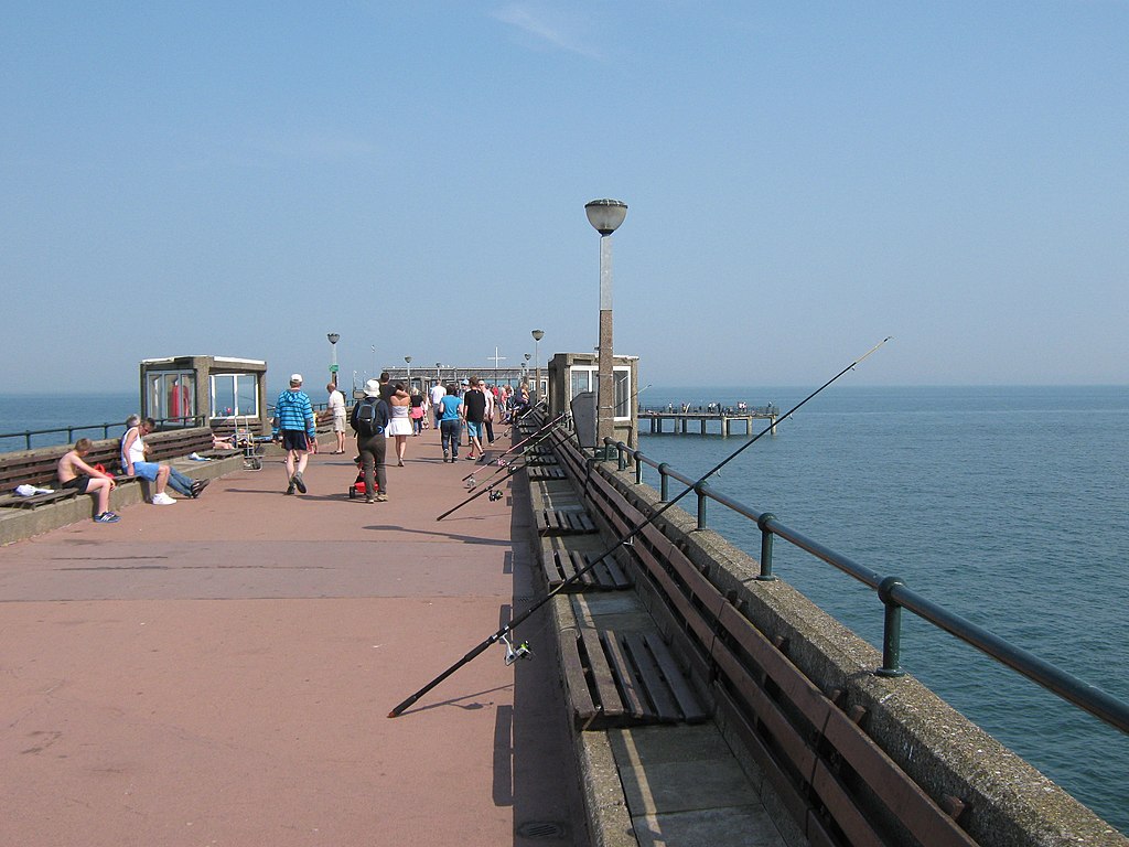 People stroll along a sunny pier with benches and fishing rods overlooking calm, blue water. The sky is clear, creating a relaxed, leisurely atmosphere.