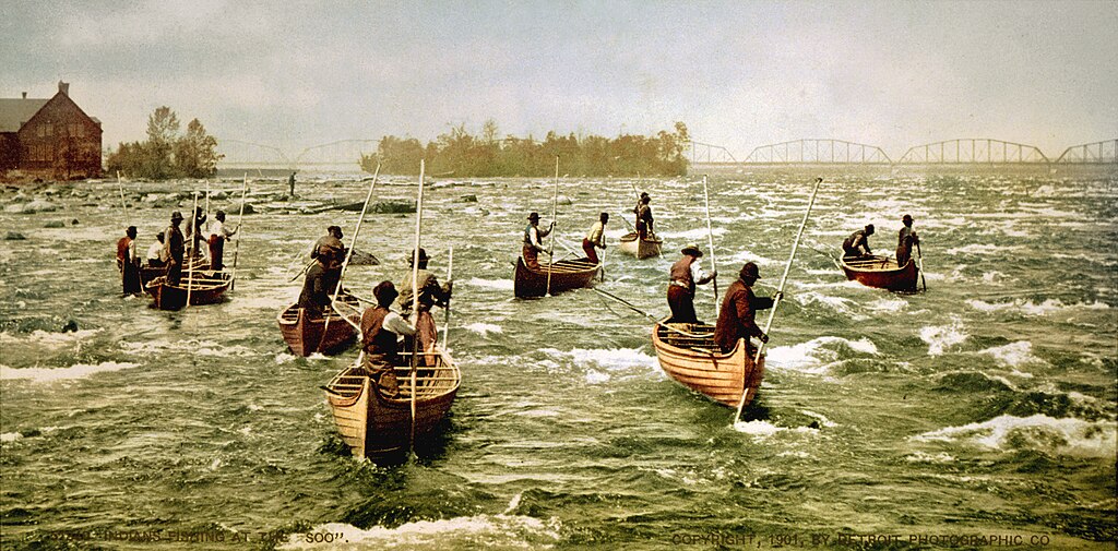 Men in small boats fish with poles in the swift currents of the St. Marys River, near the Soo Locks. A bridge and building are visible in the background.