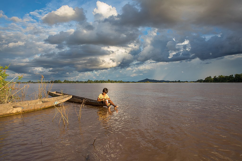 A child sits on a wooden boat in a vast, brown river under dramatic cloudy skies. The horizon shows distant green trees and a small hill.