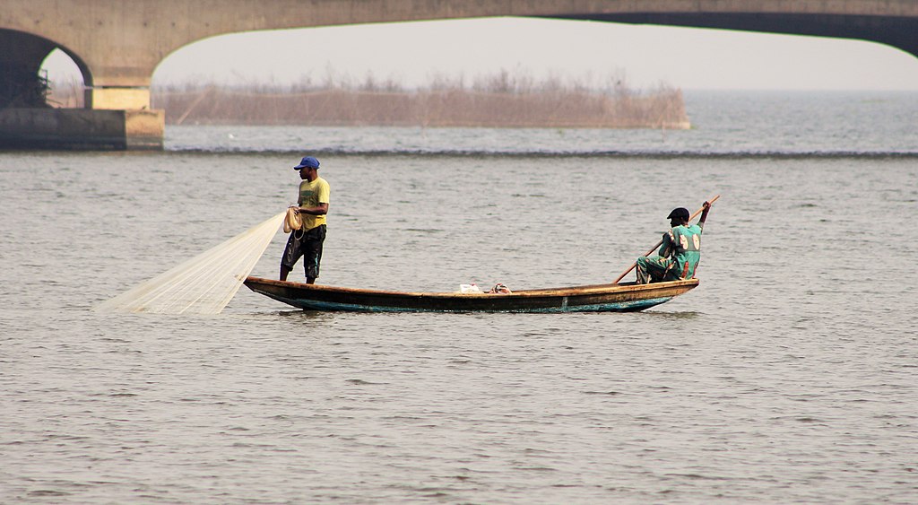 Two fishermen stand on a small wooden boat in calm waters, casting a net under a concrete bridge. The scene conveys a peaceful, focused atmosphere.