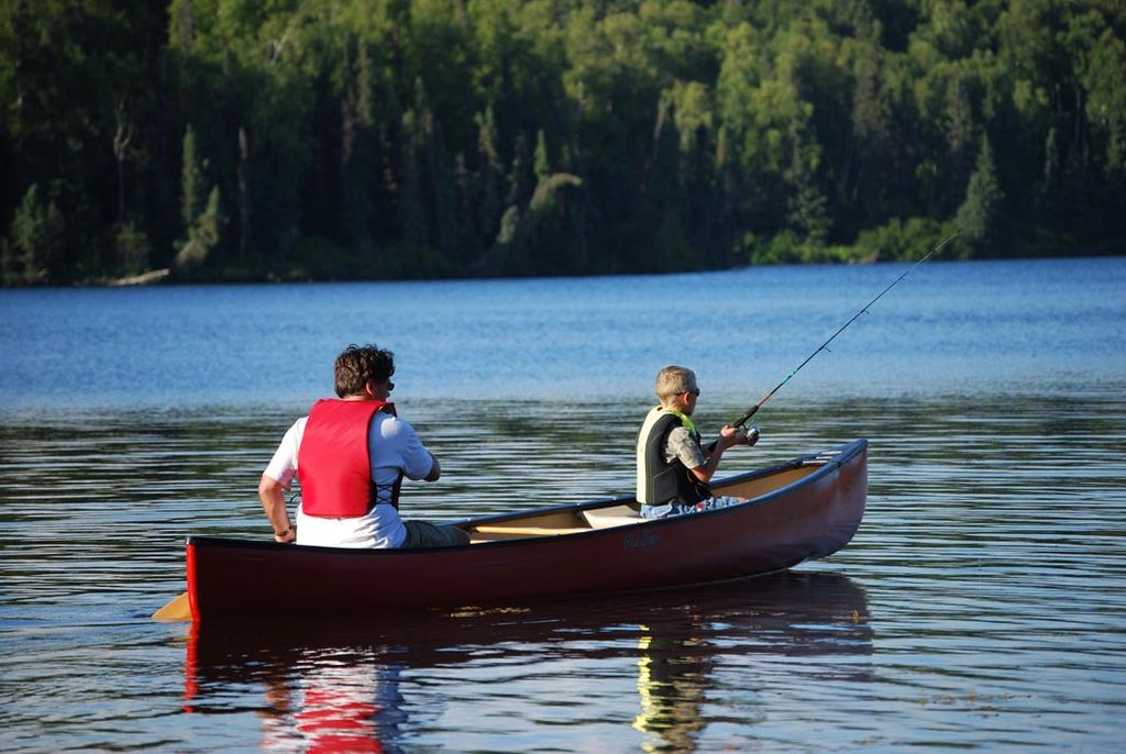 Two people fishing in a canoe on a calm lake, surrounded by lush green trees. One wears a red life jacket; the scene is peaceful and serene.