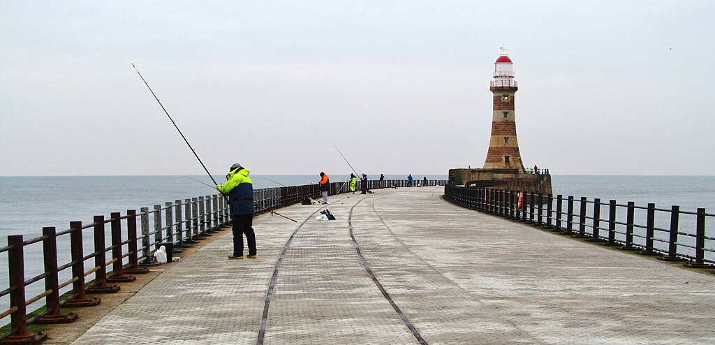 A lighthouse overlooks a long pier extending into the ocean. People are fishing along the pier, creating a peaceful scene. The sky is overcast and gray.