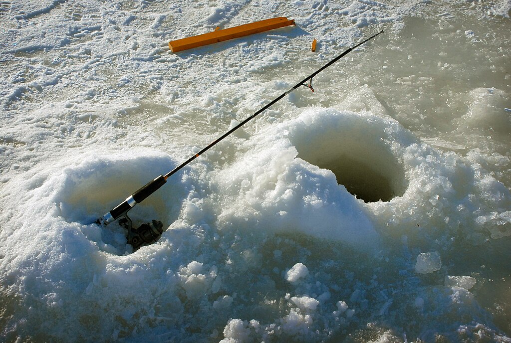 A fishing rod rests near two ice holes on a frozen lake surface. The rod leans against a snow mound, with bright sunlight reflecting off the icy landscape.