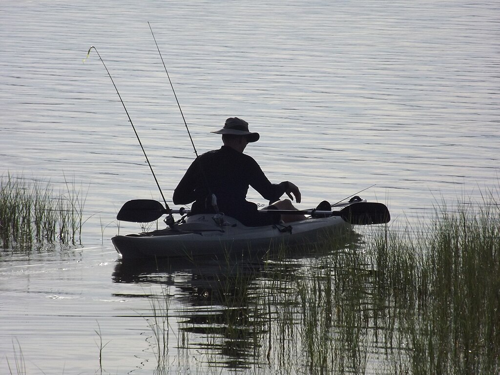 A person in a wide-brimmed hat is fishing from a kayak in calm waters, surrounded by tall grass. The scene is serene, conveying a peaceful, solitary moment.