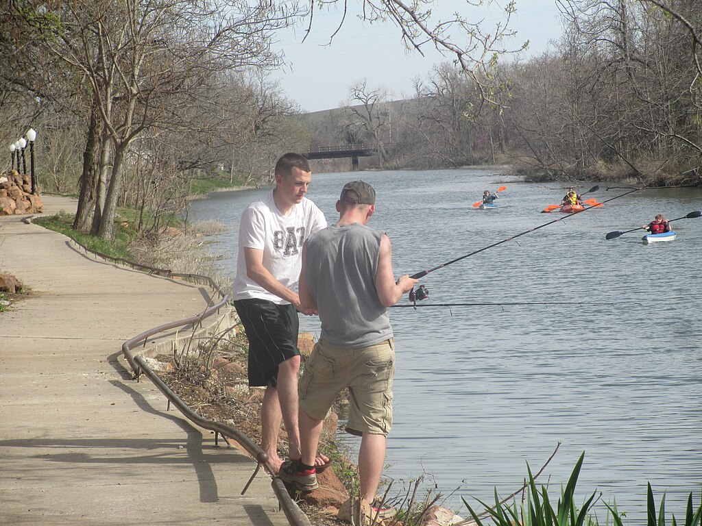 Two people fish by a calm river, standing on a concrete path with trees lining the shore. Kayakers paddle in the background, enjoying a serene day.