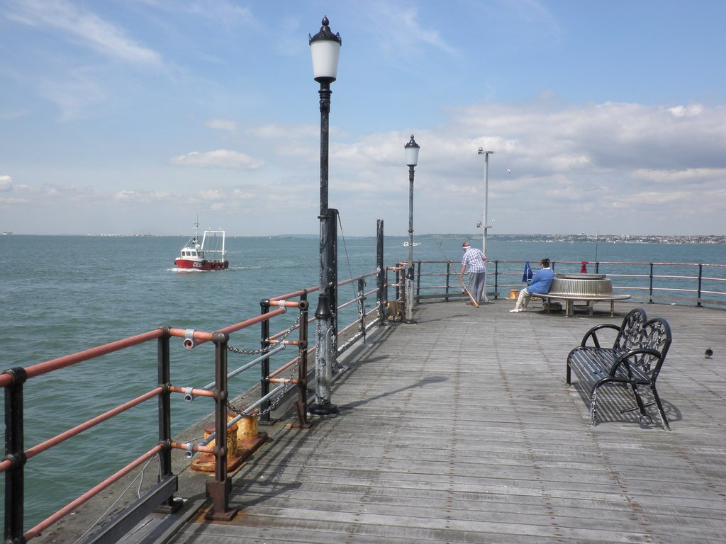 A sunny pier scene featuring a red and white boat on a calm sea. Two people are seated on a bench, with one fishing. A lamppost and sky with clouds complete the tranquil setting.