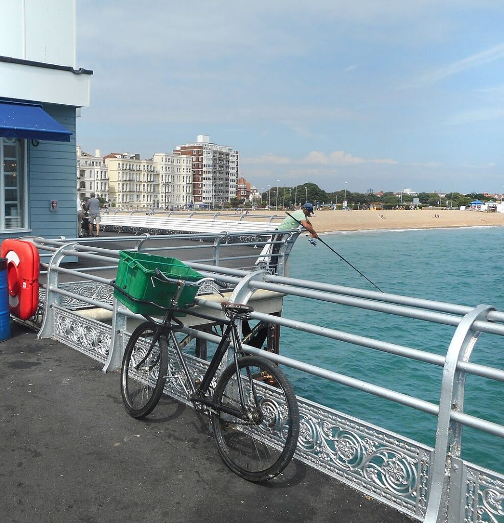 A bicycle with a green basket leans against a pier railing. A person fishes, overlooking sandy beaches and distant buildings, under a clear sky.