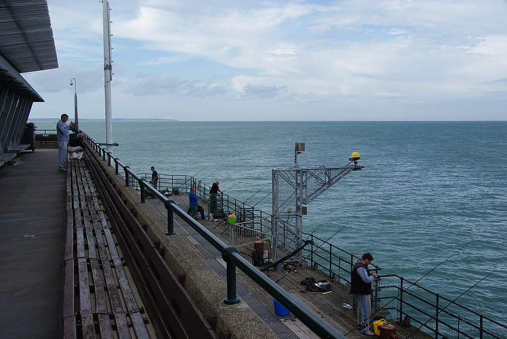 People are fishing on a pier overlooking a calm sea. The sky is overcast, and benches line the walkway. The mood is peaceful and serene.