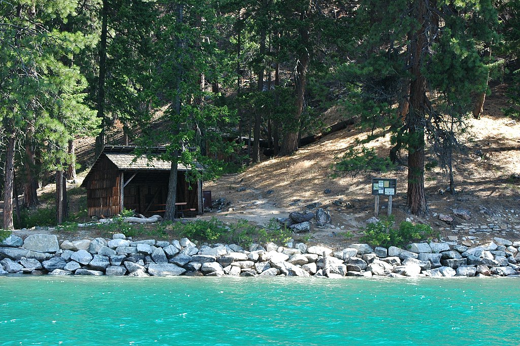 A small wooden shelted is tucked in the trees with a bright turquoise lake in the foreground. Flick Creek camp is a boat-in camp on Lake Chelan. It can also be accessed by the Lakeshore Trail.