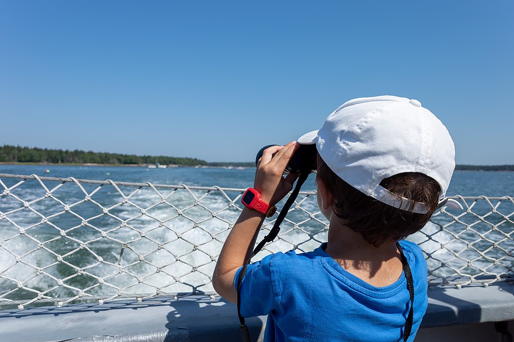 A child in a blue shirt and white cap looks through binoculars on a boat. The background shows a vibrant blue sky and ocean waves, conveying exploration.