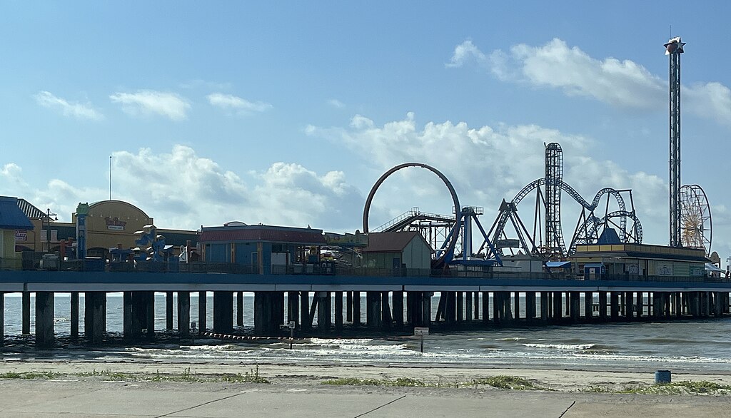 Amusement park on a pier over the ocean, featuring roller coasters and rides against a bright blue sky with clouds, creating a lively atmosphere.