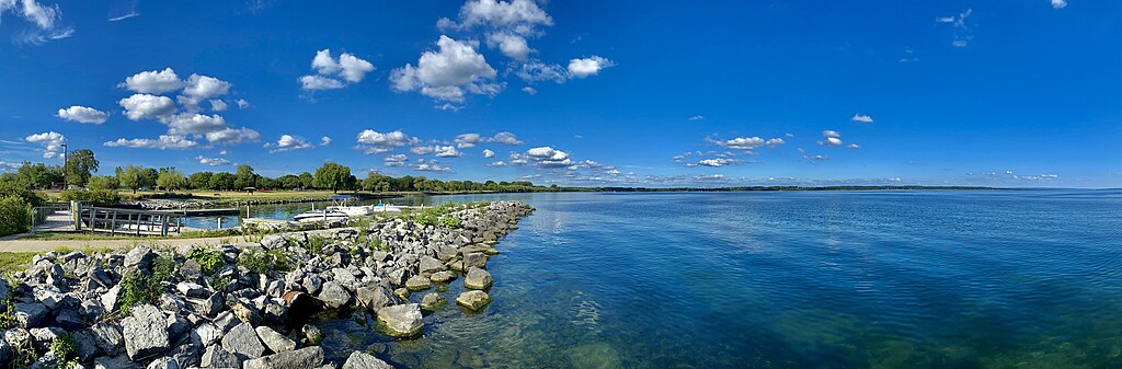 Panoramic view of a serene lake under a vibrant blue sky dotted with fluffy clouds. Rocky shoreline and green trees add a peaceful, natural feel.