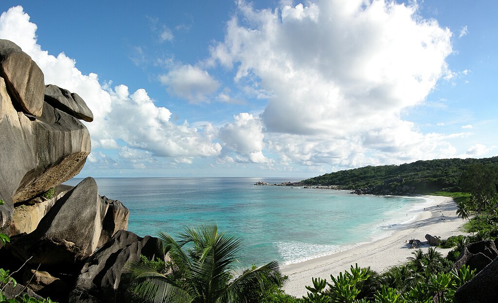 Rocky cliffs overlook a serene tropical beach with turquoise waters and white sand. Lush greenery covers the landscape under a partly cloudy blue sky.