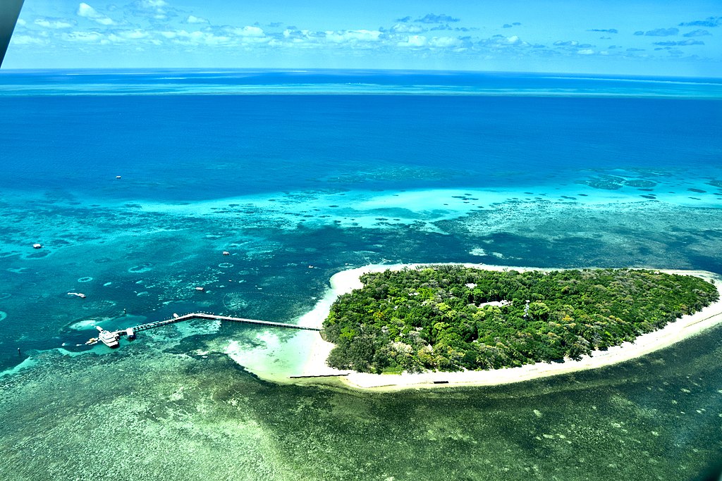 Aerial view of a lush, green island surrounded by clear blue ocean with visible coral reefs. A long pier extends into the water, evoking tranquility.