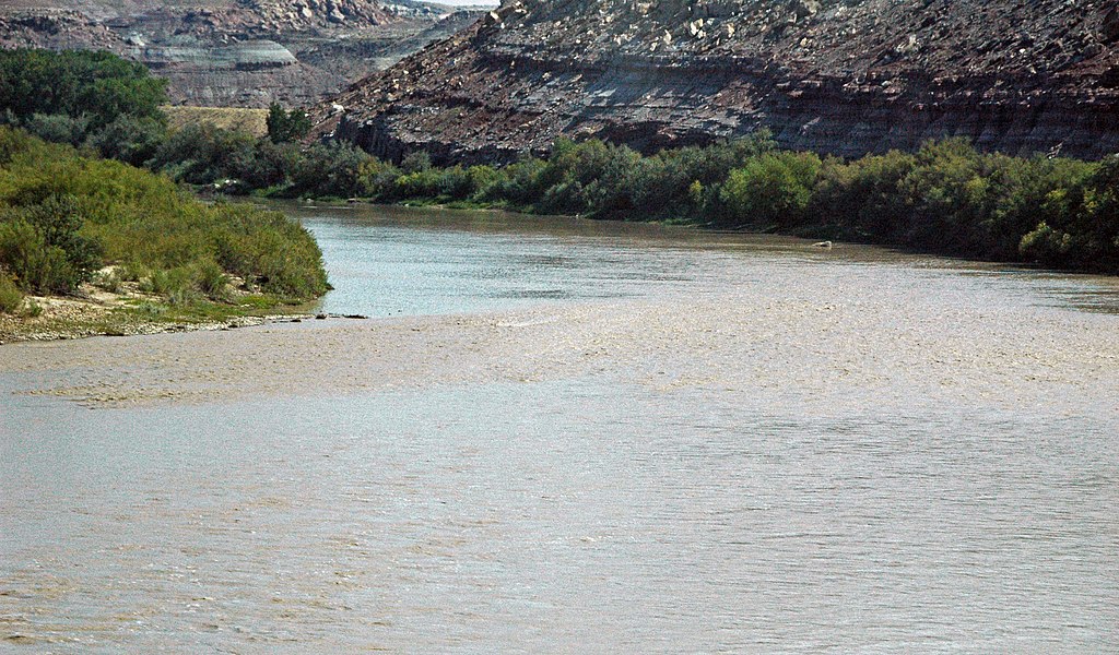 Calm river flows between green shrubs on the left and rocky cliffs on the right under a clear sky, conveying a serene, natural landscape.
