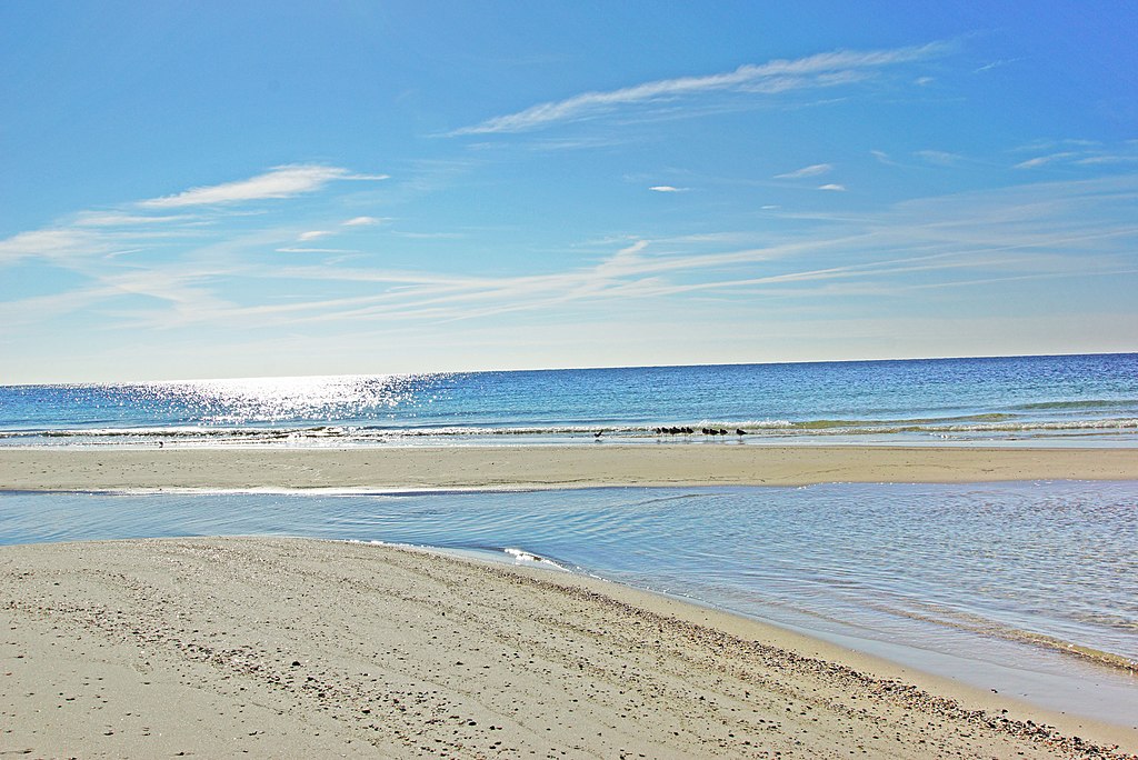 Sunny beach scene with sparkling blue waves gently lapping onto a sandy shore adorned with small ripples and a few scattered pebbles; serene and tranquil.