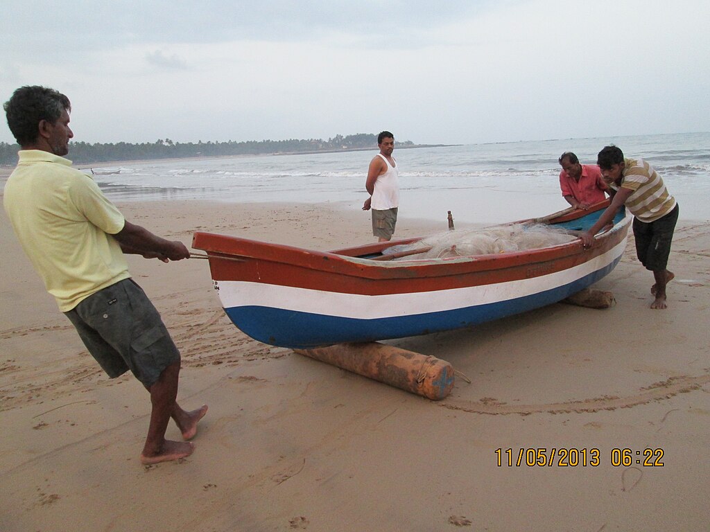 Four men push a colorful wooden boat on a sandy beach. The ocean gently waves in the background, creating a serene, collaborative atmosphere.