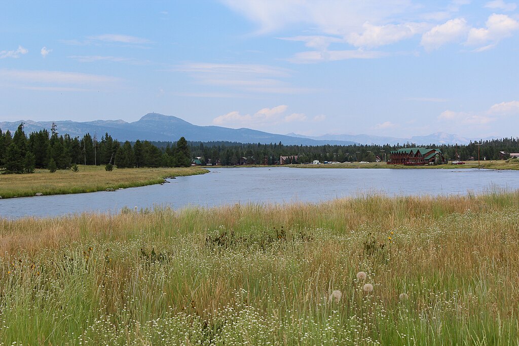 Tranquil riverside scene with lush grass in the foreground and pine trees lining the riverbank. A lodge sits near the water, mountains in the distance.