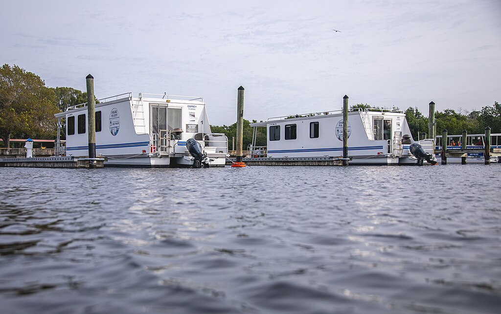 Two white houseboats docked at a marina, with a calm water surface and greenery in the background. The scene conveys a peaceful, serene ambiance.