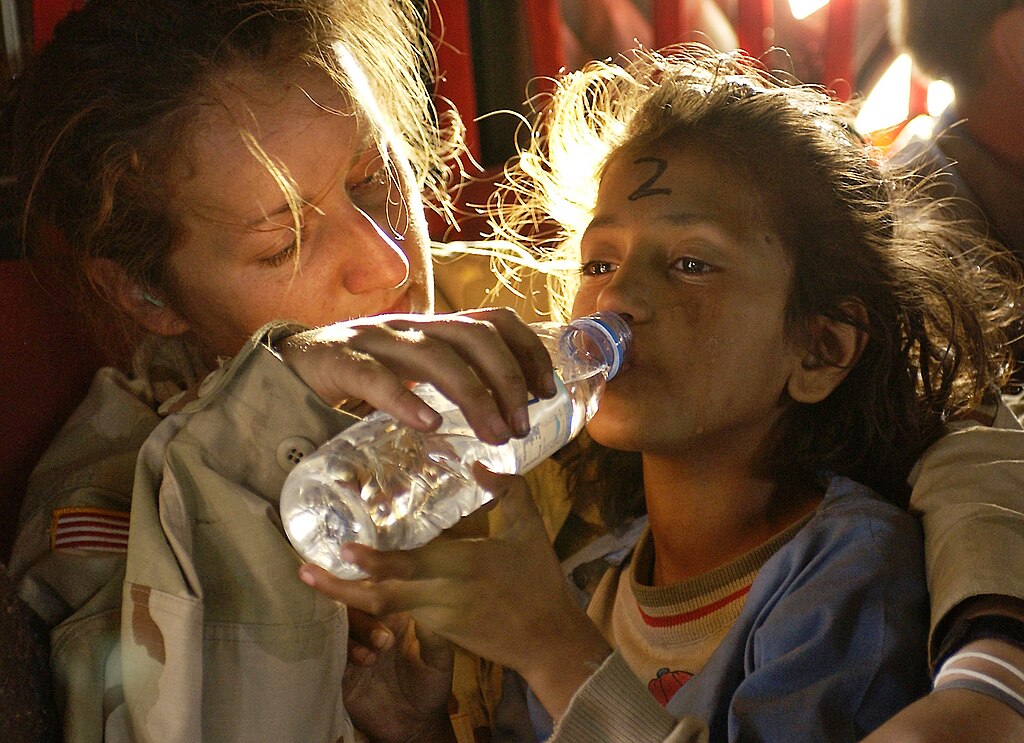 A soldier in uniform gently helps a young child drink from a water bottle. The child looks weary, with a number written on their forehead. The scene conveys compassion.