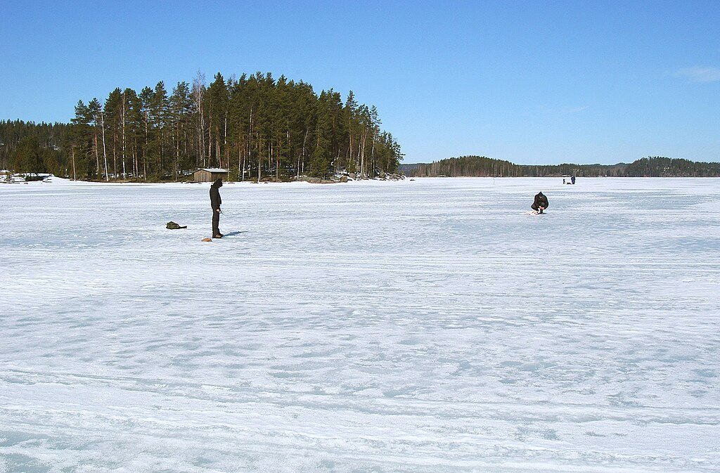 Two people are ice fishing on a frozen lake under a clear blue sky. One stands while the other kneels by a fishing hole. Snowy expanse and forested shoreline enhance the serene winter scene.