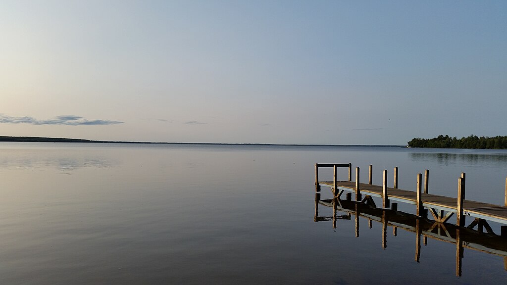 Calm lake at sunset with a wooden dock extending into the water. The sky is clear with soft pastel hues. Trees line the horizon, creating a peaceful scene.