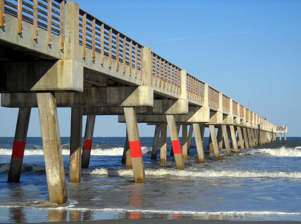 Concrete pier extending over a calm ocean, supported by pillars with red markings. Waves gently crash, under a clear blue sky. Tranquil mood.