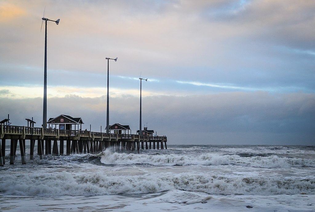 A windswept pier stretches into a rough ocean, with waves crashing below. Wind turbines stand tall against a moody sky, evoking a sense of stormy tranquility.