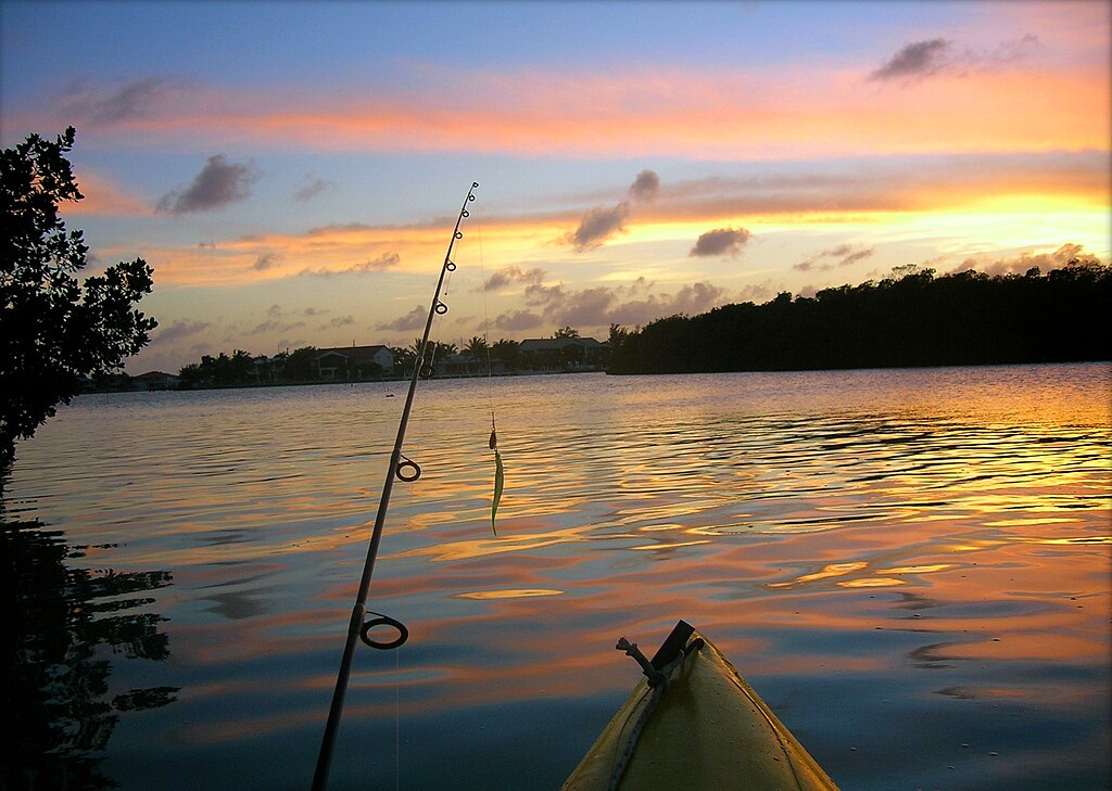Sunset over a calm lake with a fishing rod visible from a kayak. The sky is painted in soft oranges and purples, creating a peaceful, serene atmosphere.
