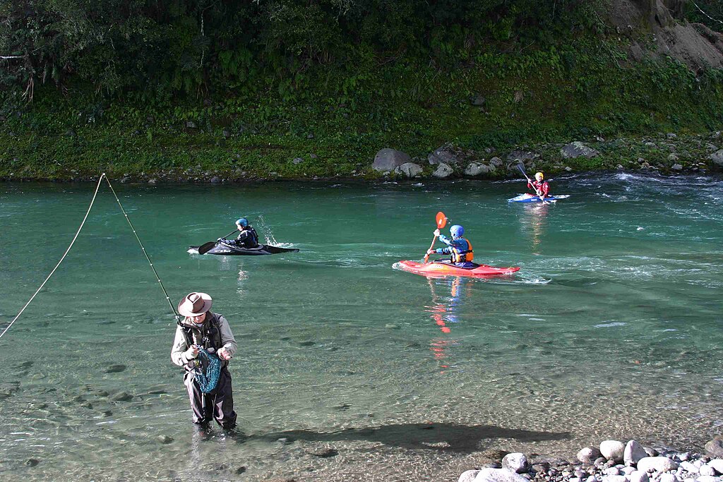 A serene river scene shows a fisherman with a net in the foreground, while three kayakers paddle through clear, green water amidst a lush, forested backdrop.