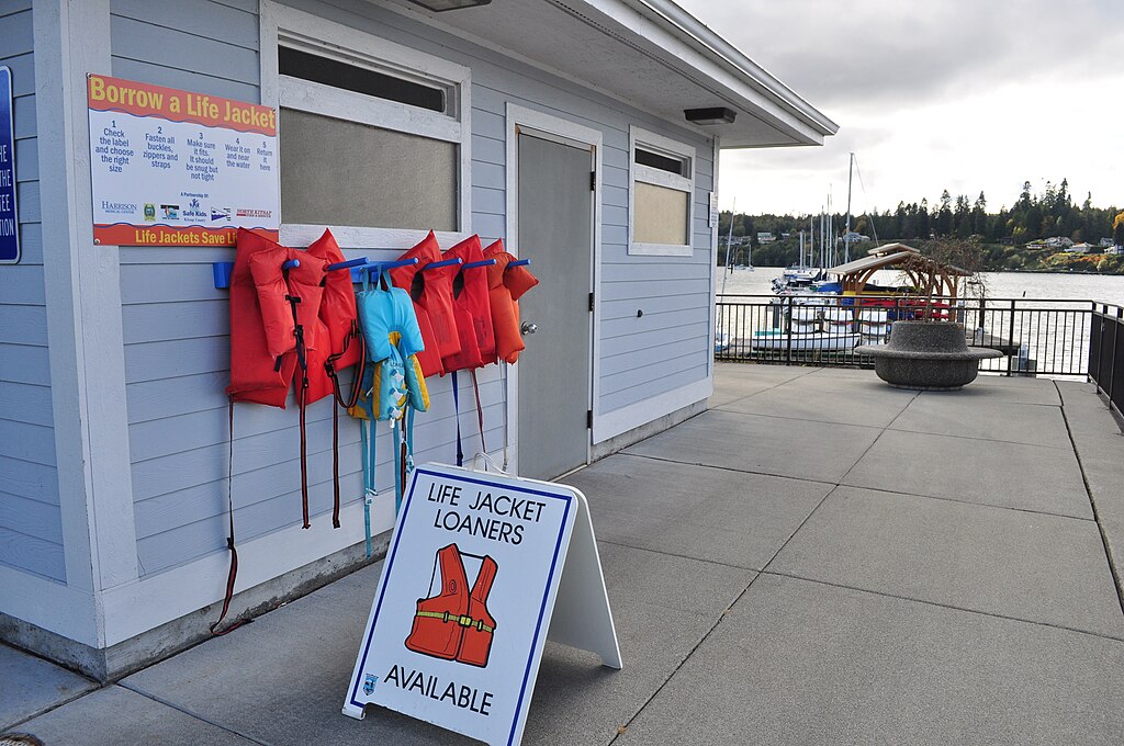 Outdoor life jacket loan station near a waterfront. Red and blue life jackets hang on a blue building. A sign reads, "Life jacket loaners available."