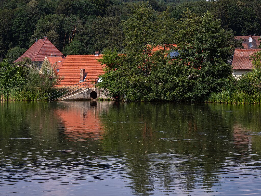 A tranquil pond reflects red-roofed houses partially hidden by lush greenery. A small concrete structure with an arch is visible by the water's edge.