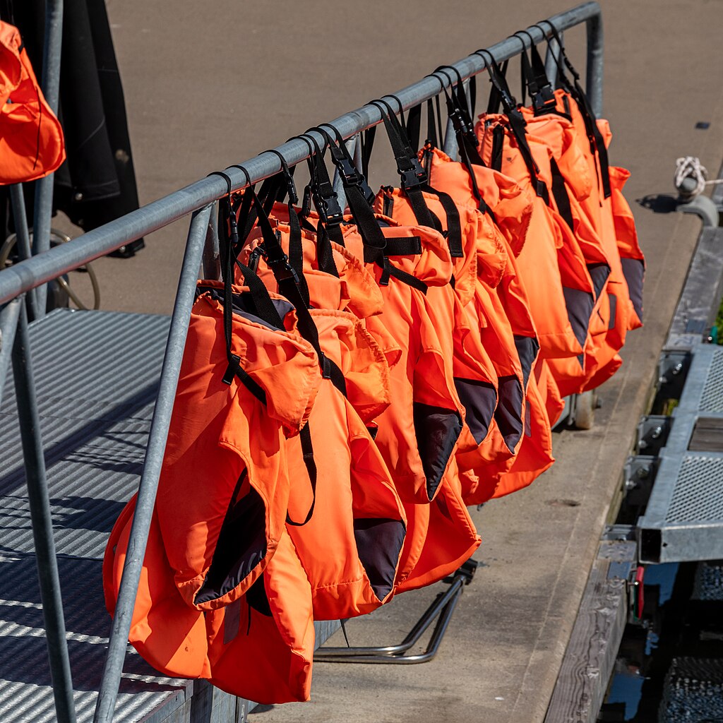 Bright orange life jackets hang neatly in a row on a metal railing. Sunlight casts shadows, creating a sense of order and readiness by the water.