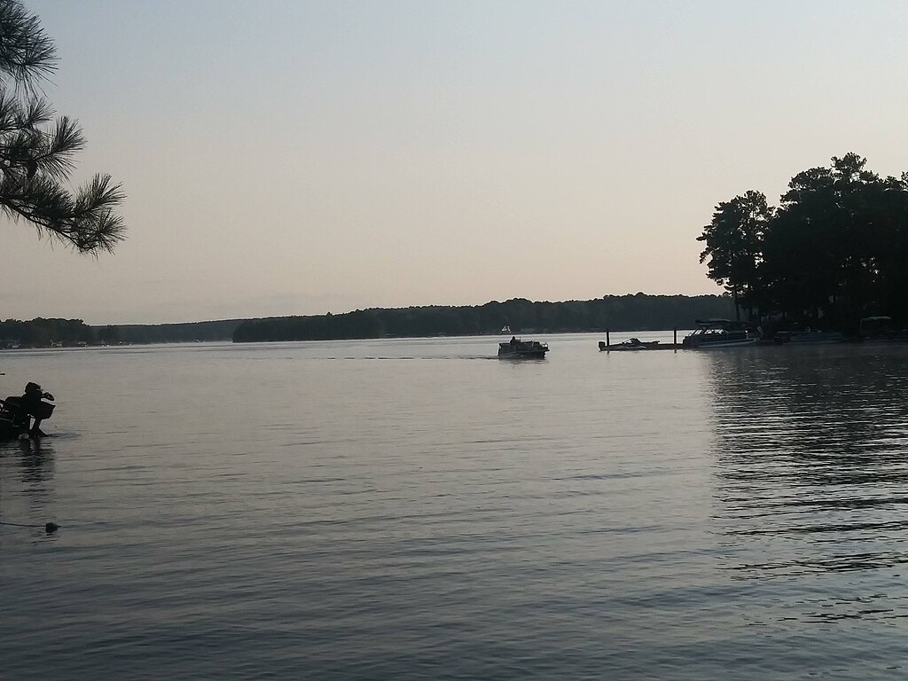 Calm lake scenery at dusk, featuring a boat moving across the water, with silhouetted trees on the shore and a sky in soft pastel tones.