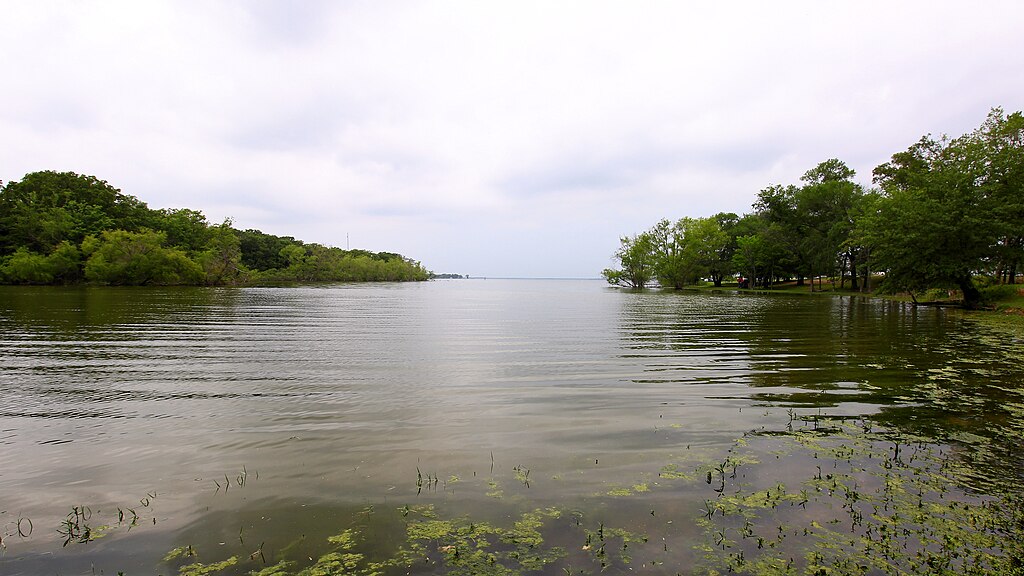 Calm lake with rippling water reflecting a cloudy sky. Lush greenery lines both shores, creating a serene and peaceful natural scene.