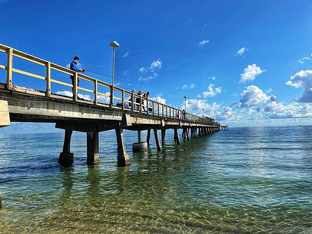 A sunny pier extends into the ocean, with people fishing and relaxing. The sky is bright blue, dotted with fluffy clouds, creating a serene atmosphere.