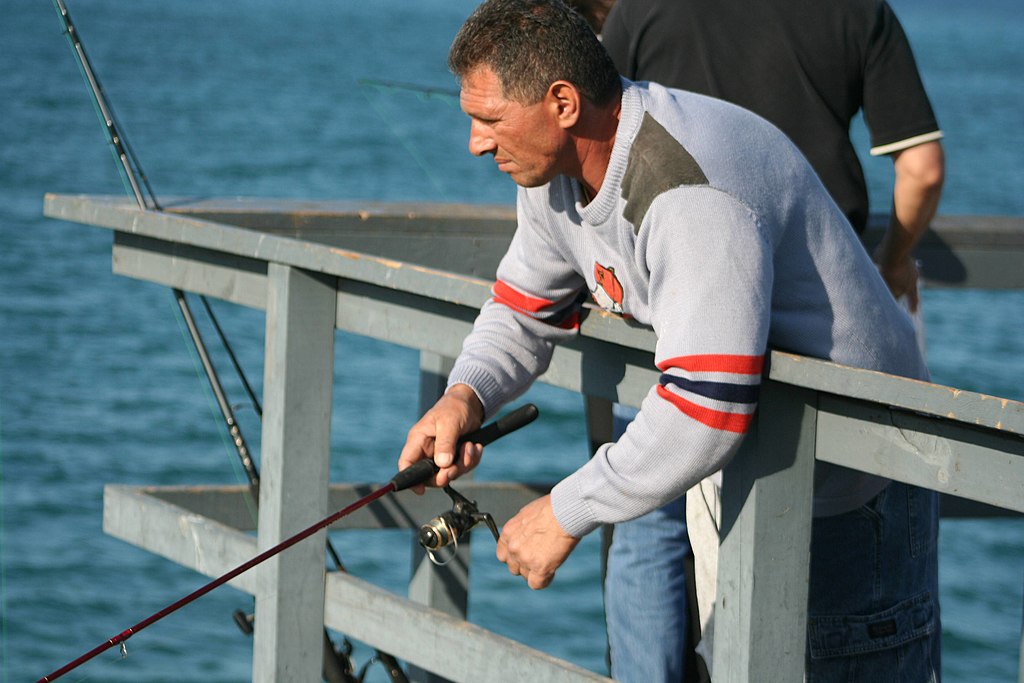 A man in a gray sweater focused on fishing from a pier overlooking blue water. He holds a fishing rod, embodying concentration and calm.