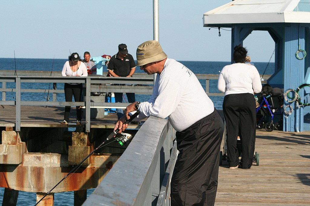 Man in a beige hat fishing from a pier, leaning over a gray railing. Several people in casual attire in the background, with the ocean visible. Calm and relaxed atmosphere.