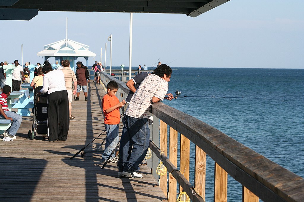 People on a sunny pier: a man fishes over railing, an orange-shirted child nearby. Others stroll, enjoy the ocean view. Relaxed, social atmosphere.